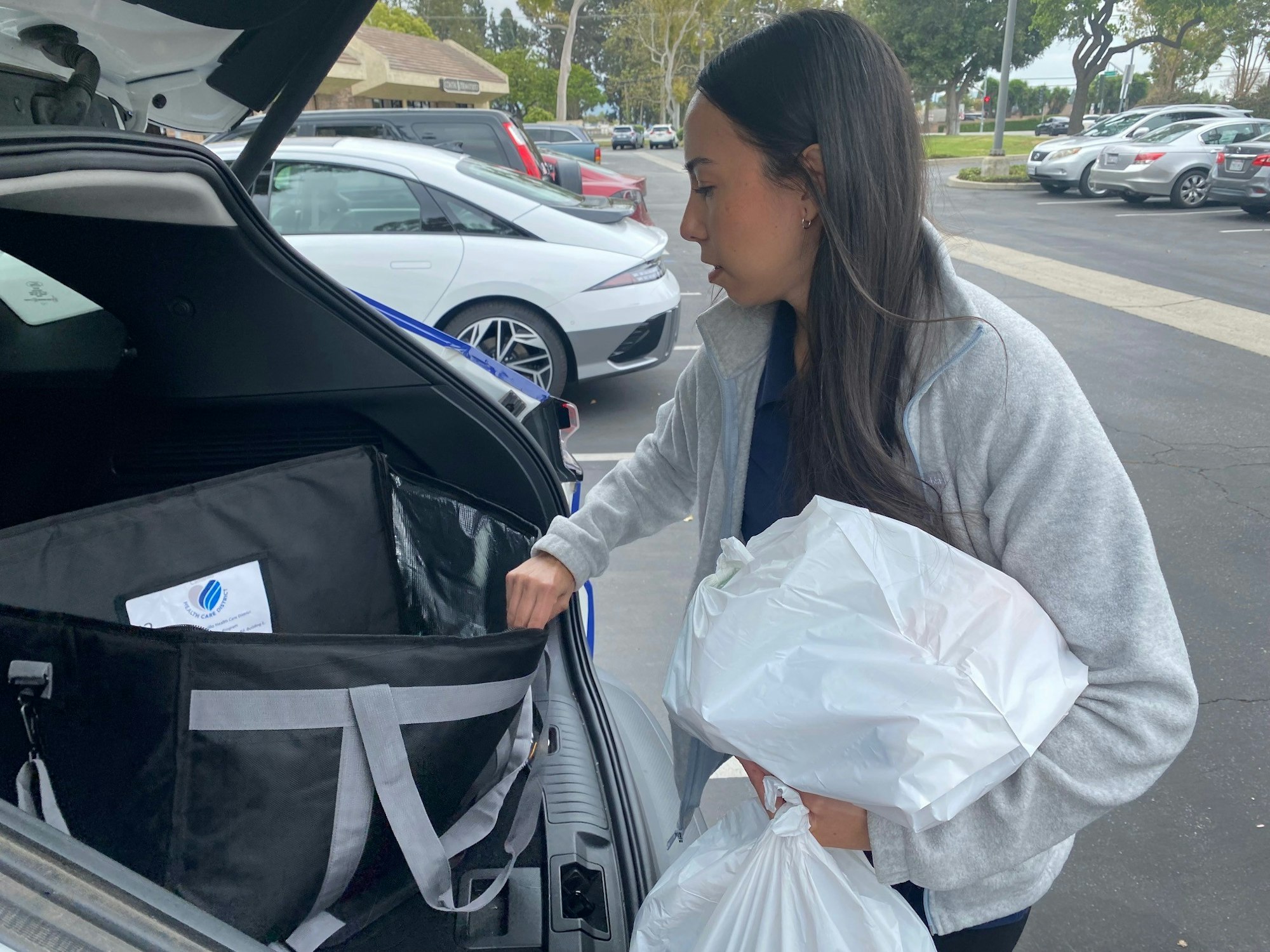 A woman is loading a large insulated delivery bag into a car trunk while holding plastic bags.