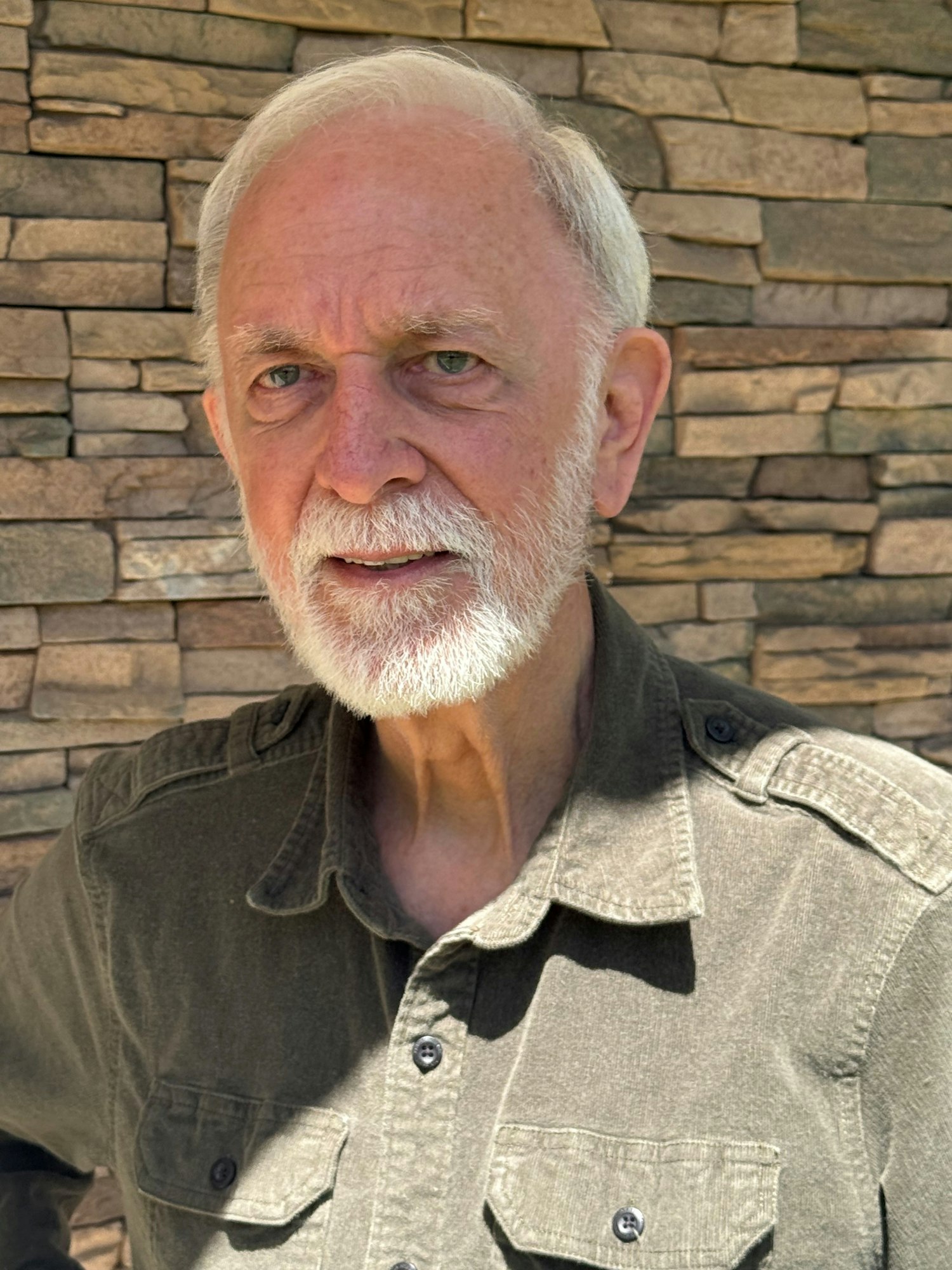 An older man with a white beard and gray hair, wearing a dark green button-up shirt, stands in front of a stone wall.
