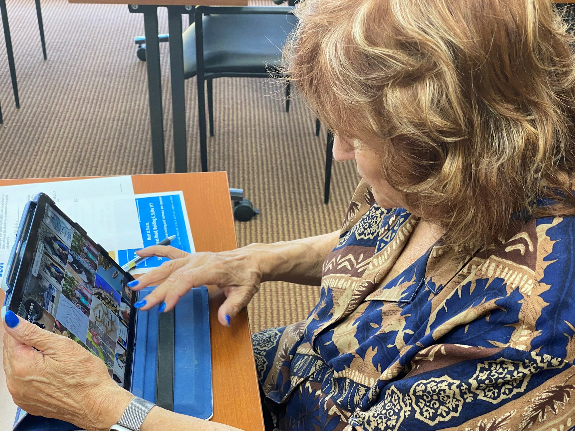 A person with painted nails uses a tablet, browsing photos, while seated at a table with papers and a pen nearby.