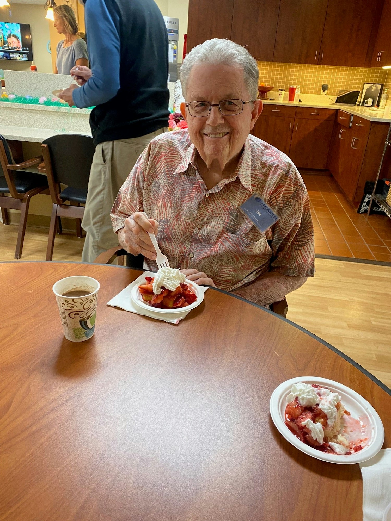 A person sitting at a table, smiling, with strawberry shortcake and coffee. Other people in the kitchen background.