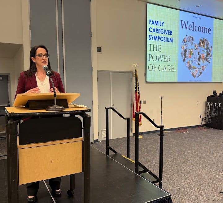 A speaker presents at the Family Caregiver Symposium, discussing the theme "The Power of Care" with a welcoming backdrop.