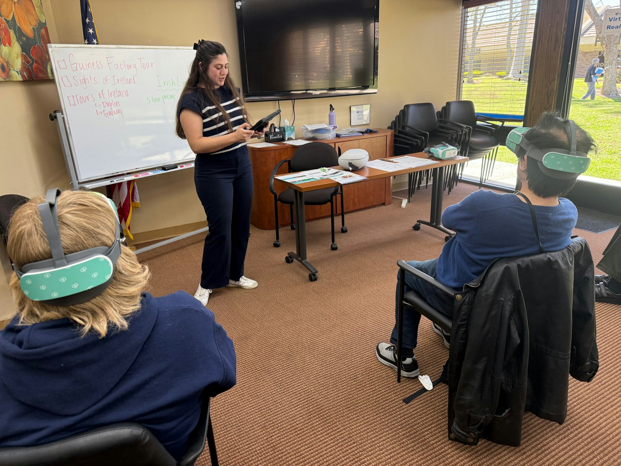 A woman presents in a room; attendees with VR headsets are seated, whiteboard lists Ireland tour items.