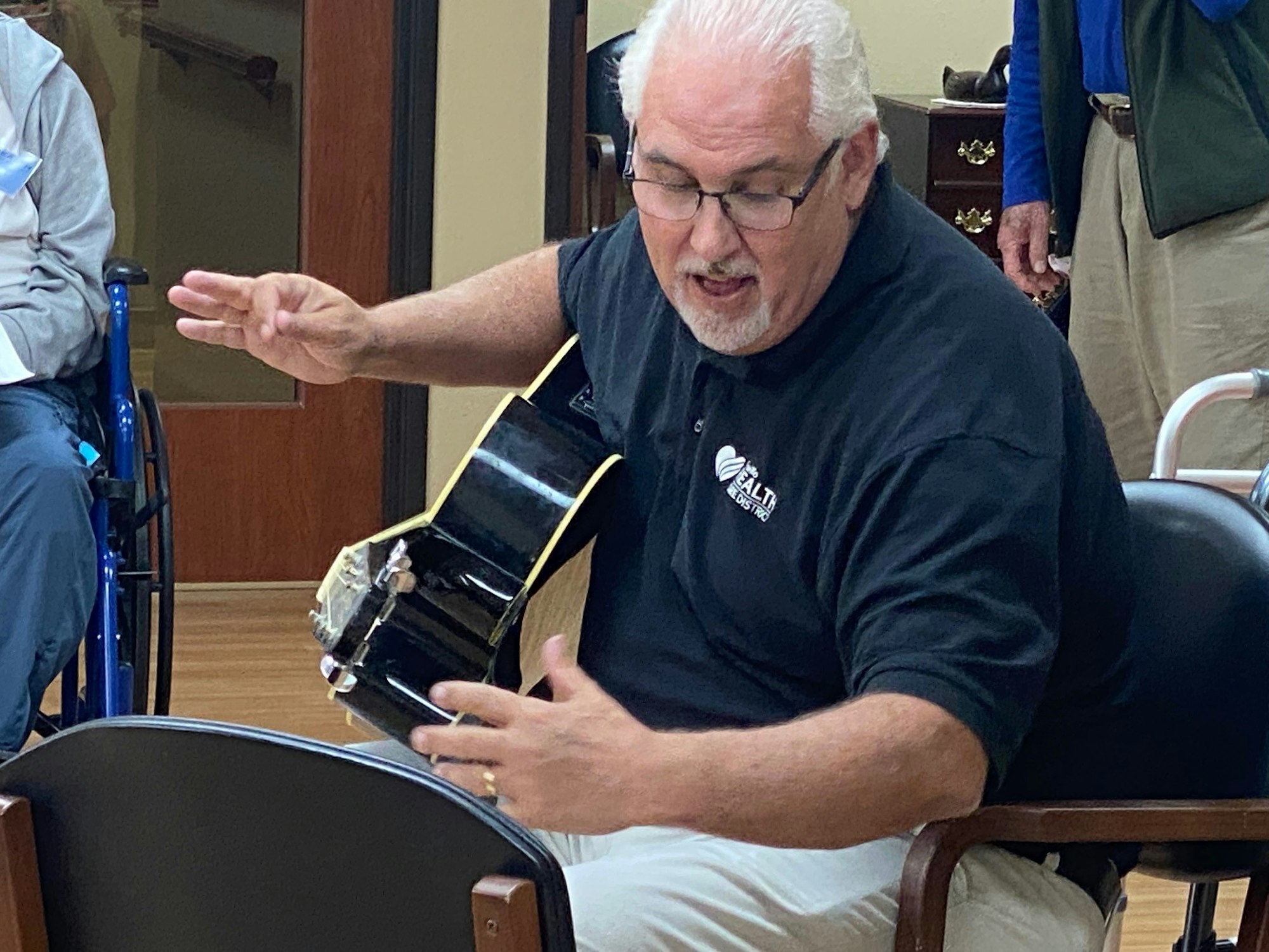 Man with a guitar gesturing, wearing a black shirt with a logo, indoors.