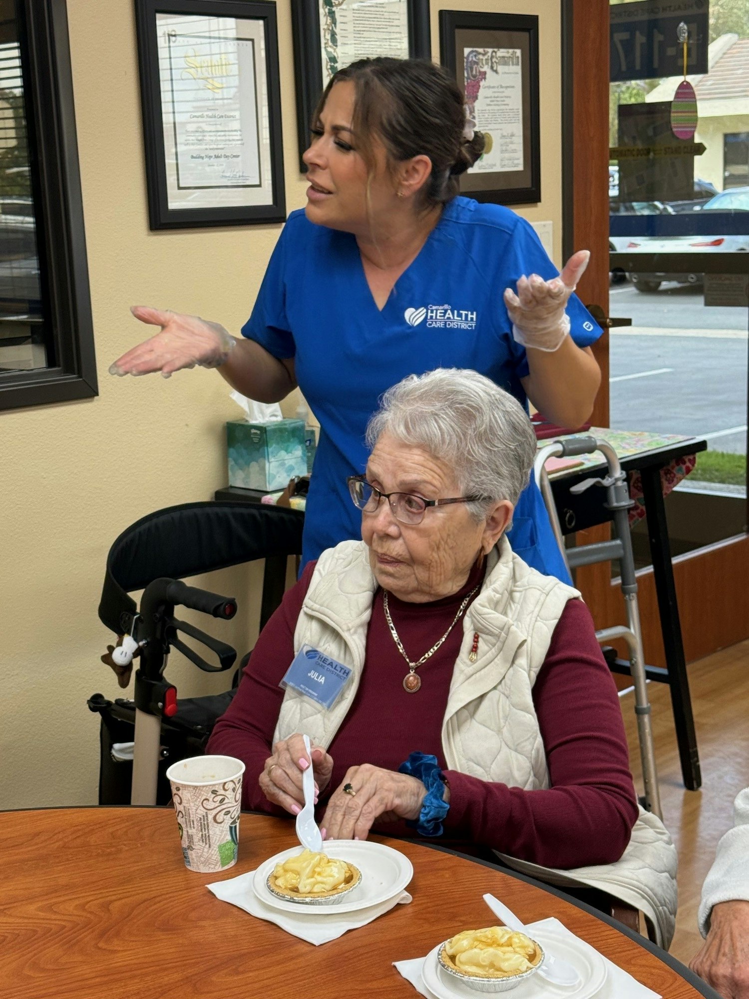 A caregiver in scrubs appears to be expressing concern while an elderly woman enjoys dessert at a table.