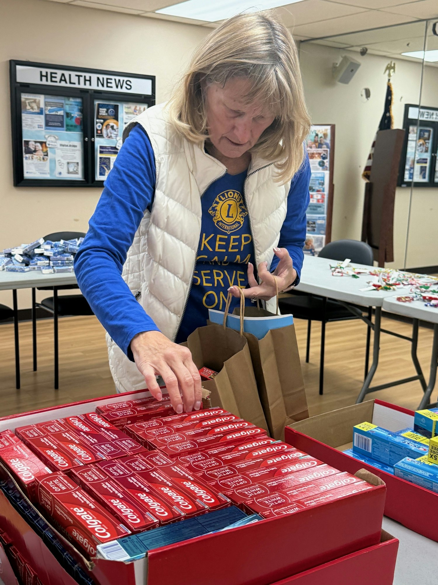 A woman packs boxes of Colgate toothpaste into a bag at a community center with health news on the wall.