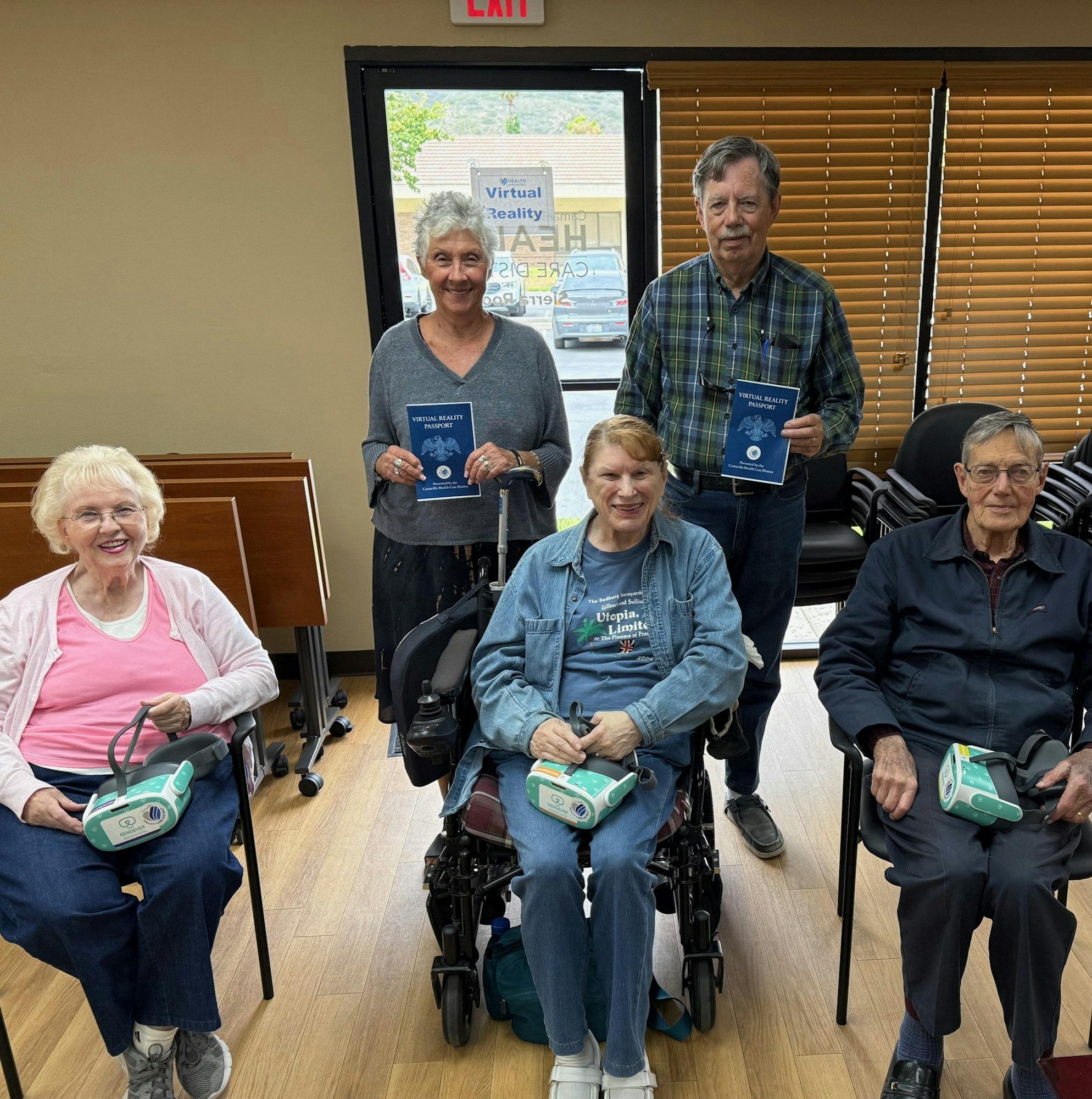 Group of seniors smiling, some holding virtual reality brochures, others holding VR headsets, sitting and standing in a room.