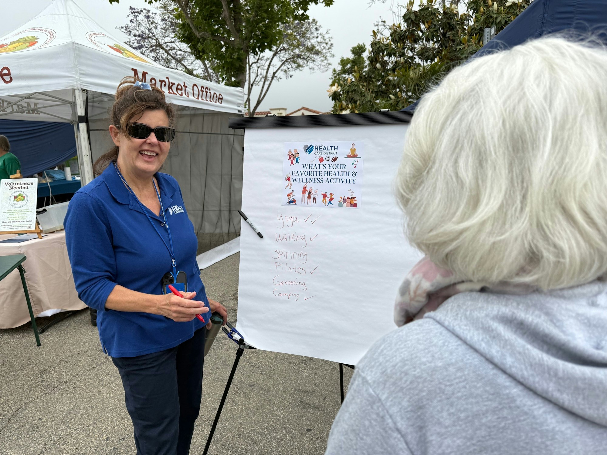 Two women at an outdoor event discuss health and wellness activities listed on a board.