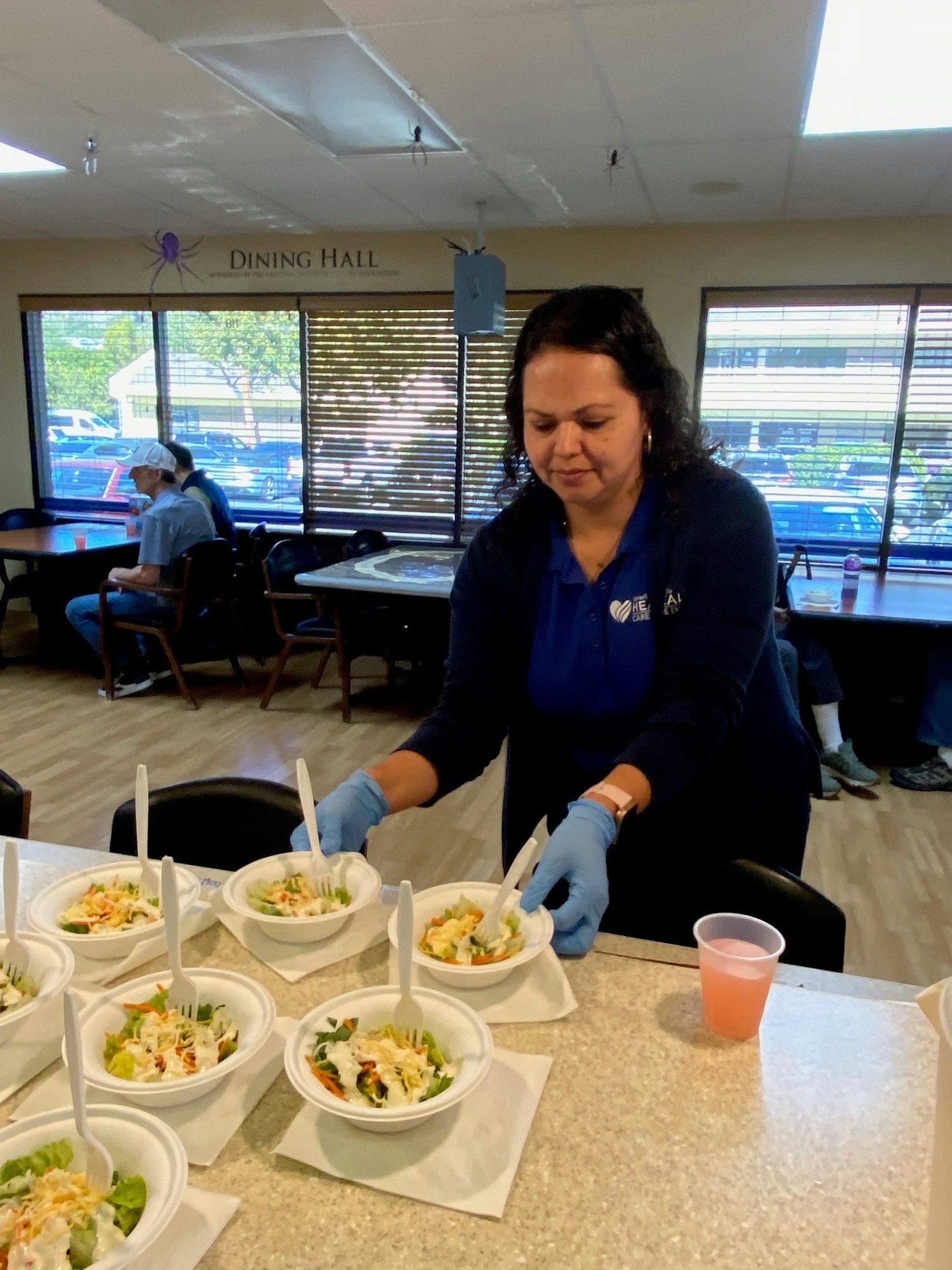 A person is preparing multiple salad bowls in a dining hall, wearing blue gloves. There are people in the background.