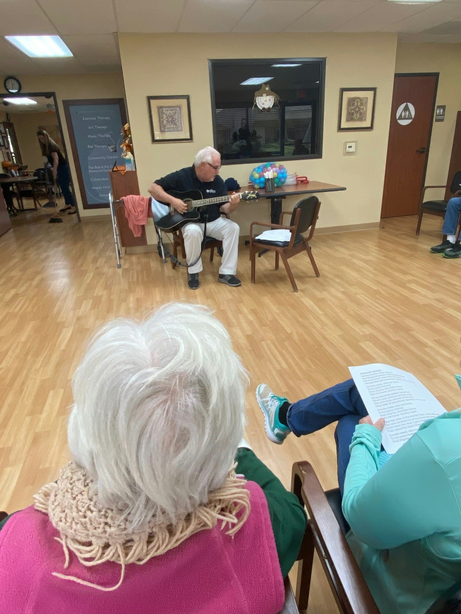A man plays guitar for an audience in a room, with a listener holding a sheet of paper in the foreground.
