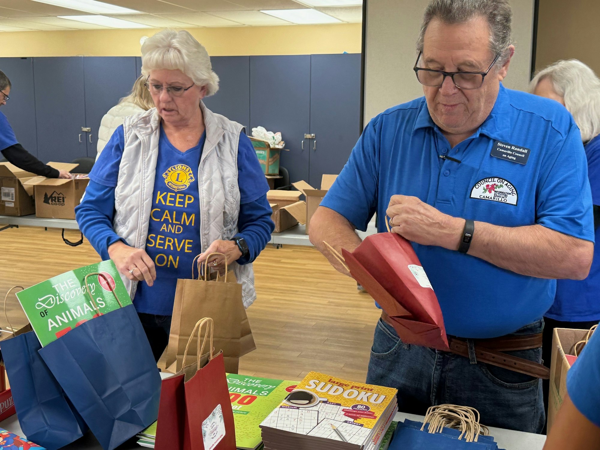 Two people are assembling gift bags with books and puzzles, wearing volunteer shirts.