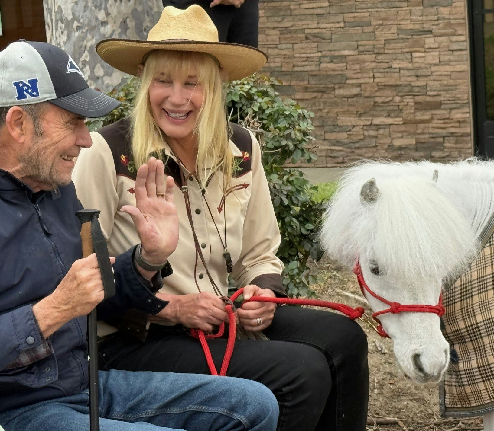 A man and woman smiling with a small white pony on a leash outdoors.