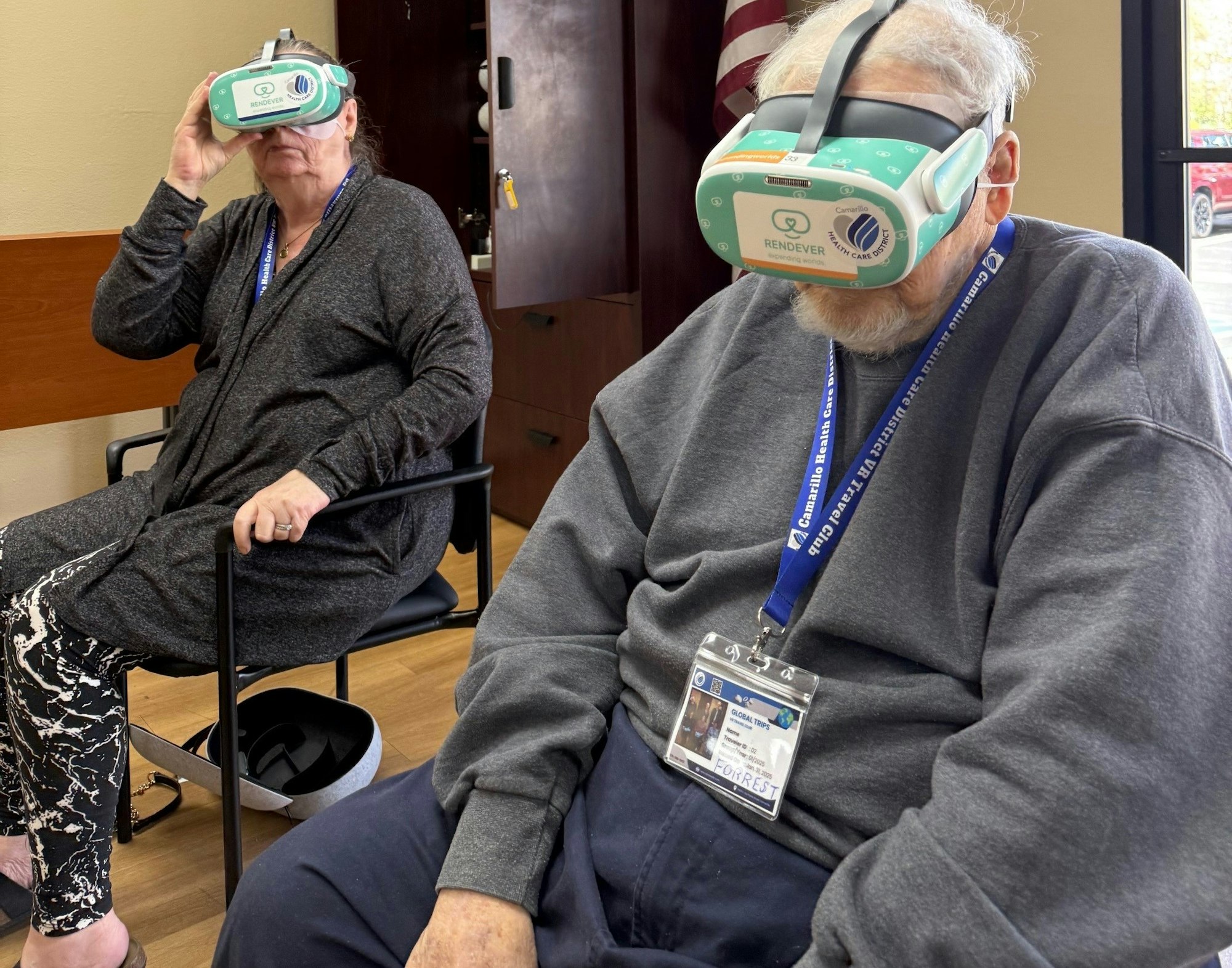 Two people seated indoors wearing VR headsets, with lanyards around their necks.