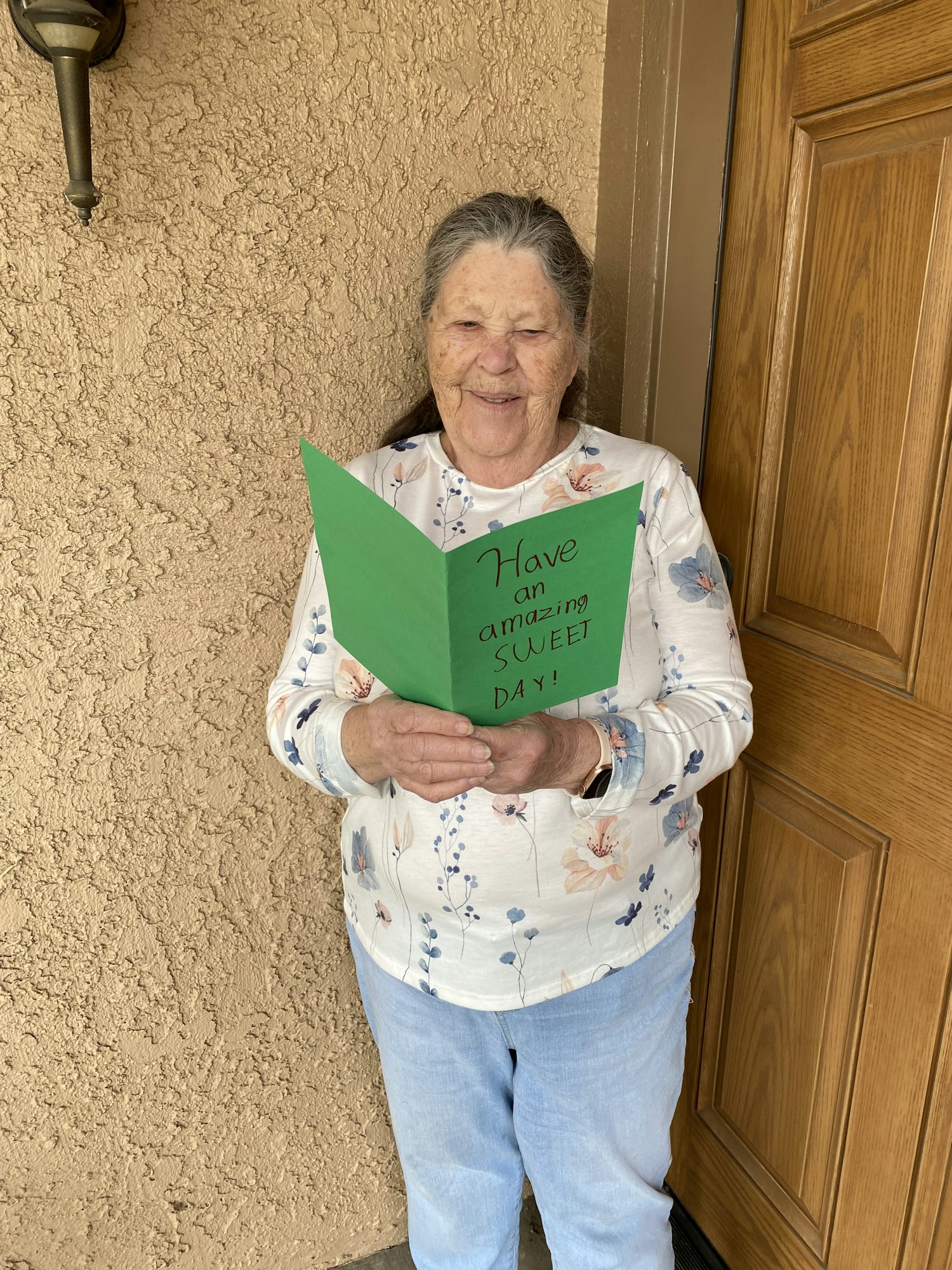 An elderly woman smiling, holding a green card that says, "Have an Amazing Day!"