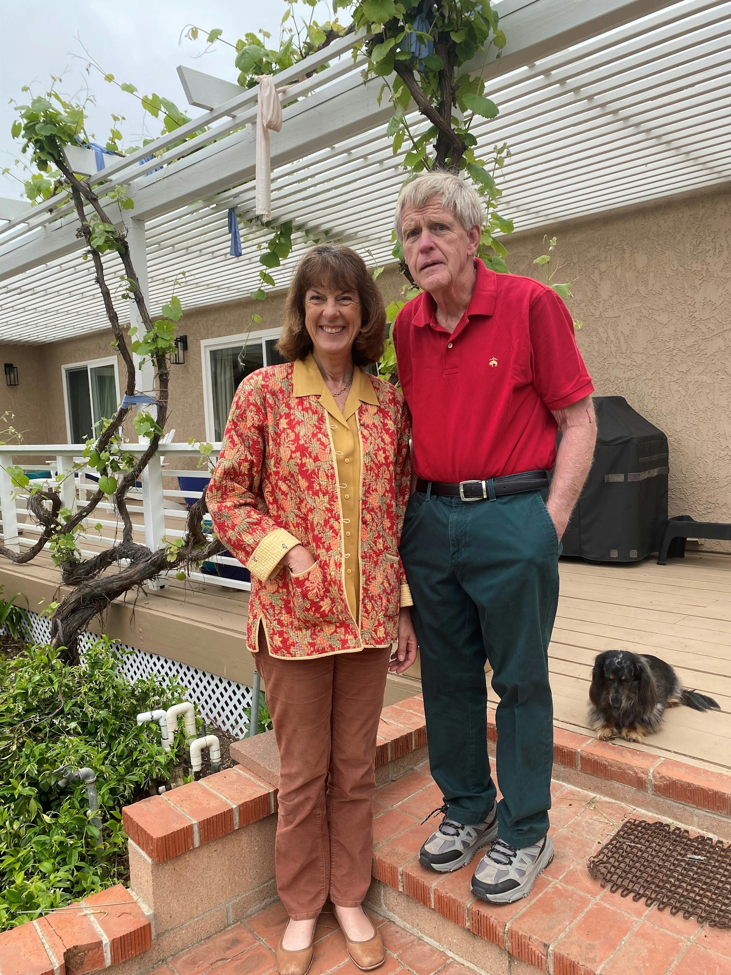 Two smiling people posing outside by a house with a pergola and a small dog in the background.