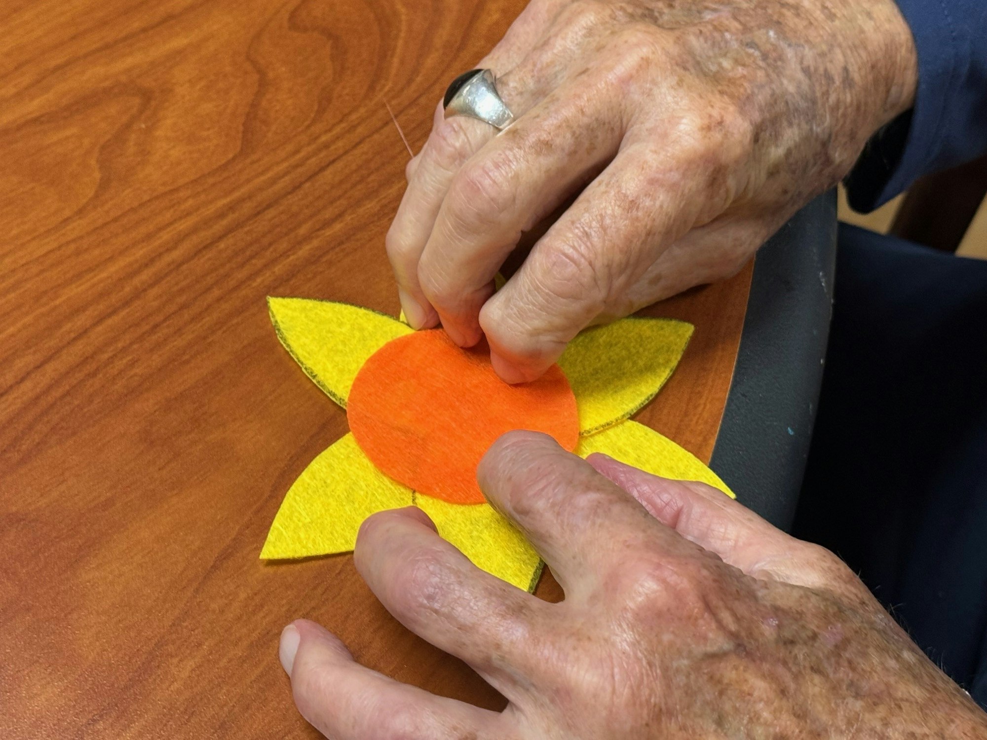 Hands crafting a felt flower with yellow petals and an orange center on a wooden table.