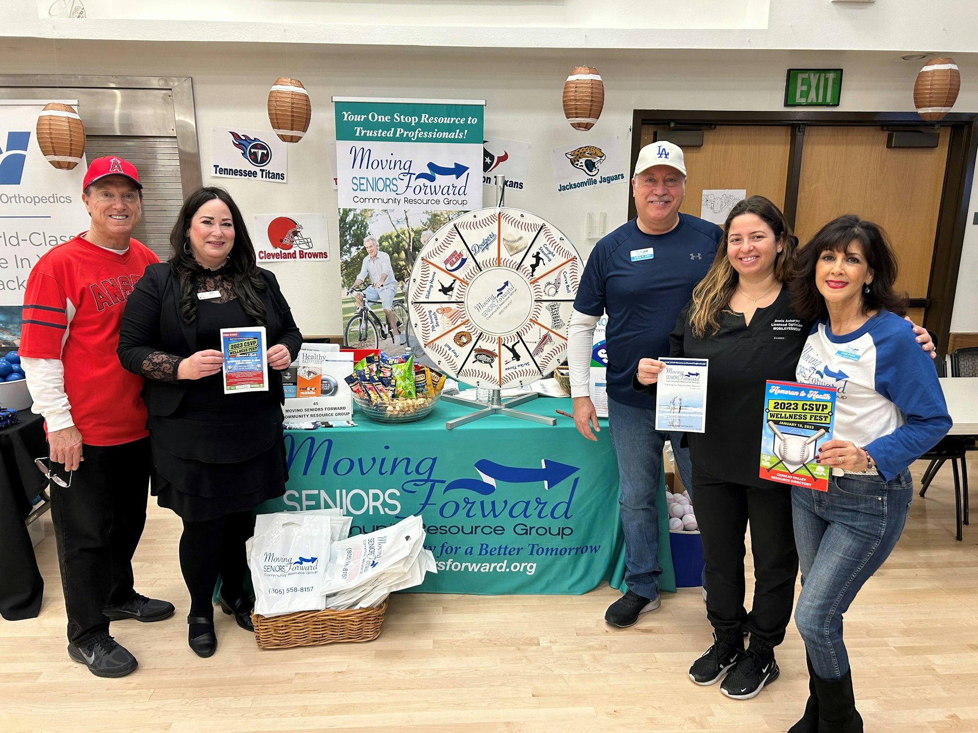Five people stand smiling by a booth with banners, sports memorabilia, and a prize wheel, at a community event.