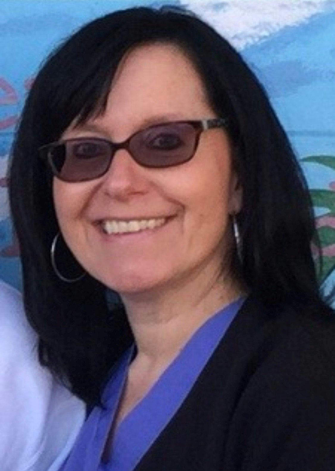 A smiling woman with dark hair and sunglasses, wearing a blue top and black jacket.