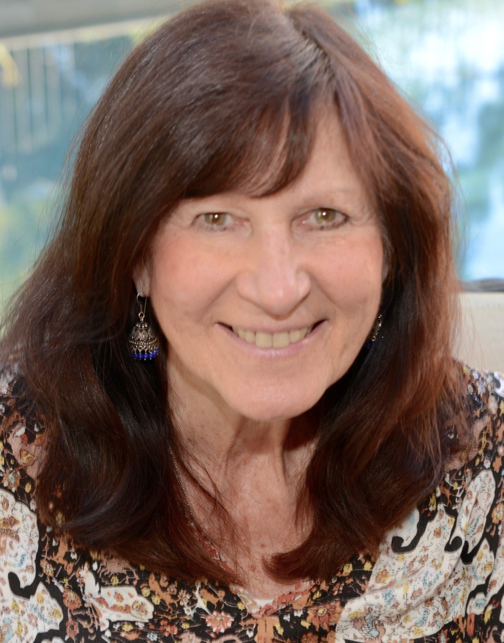 A smiling woman with long hair wearing patterned clothes and earrings against a blurred background.