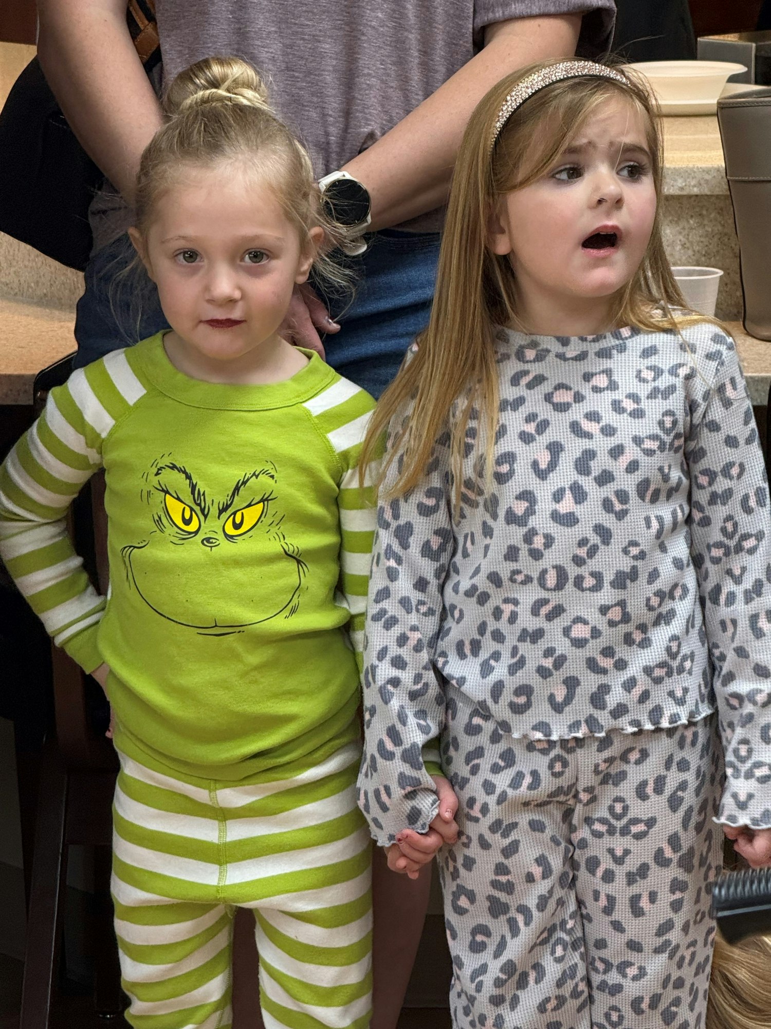Two children in pajamas: one in a Grinch-themed set, the other in leopard print, holding hands in a kitchen setting.