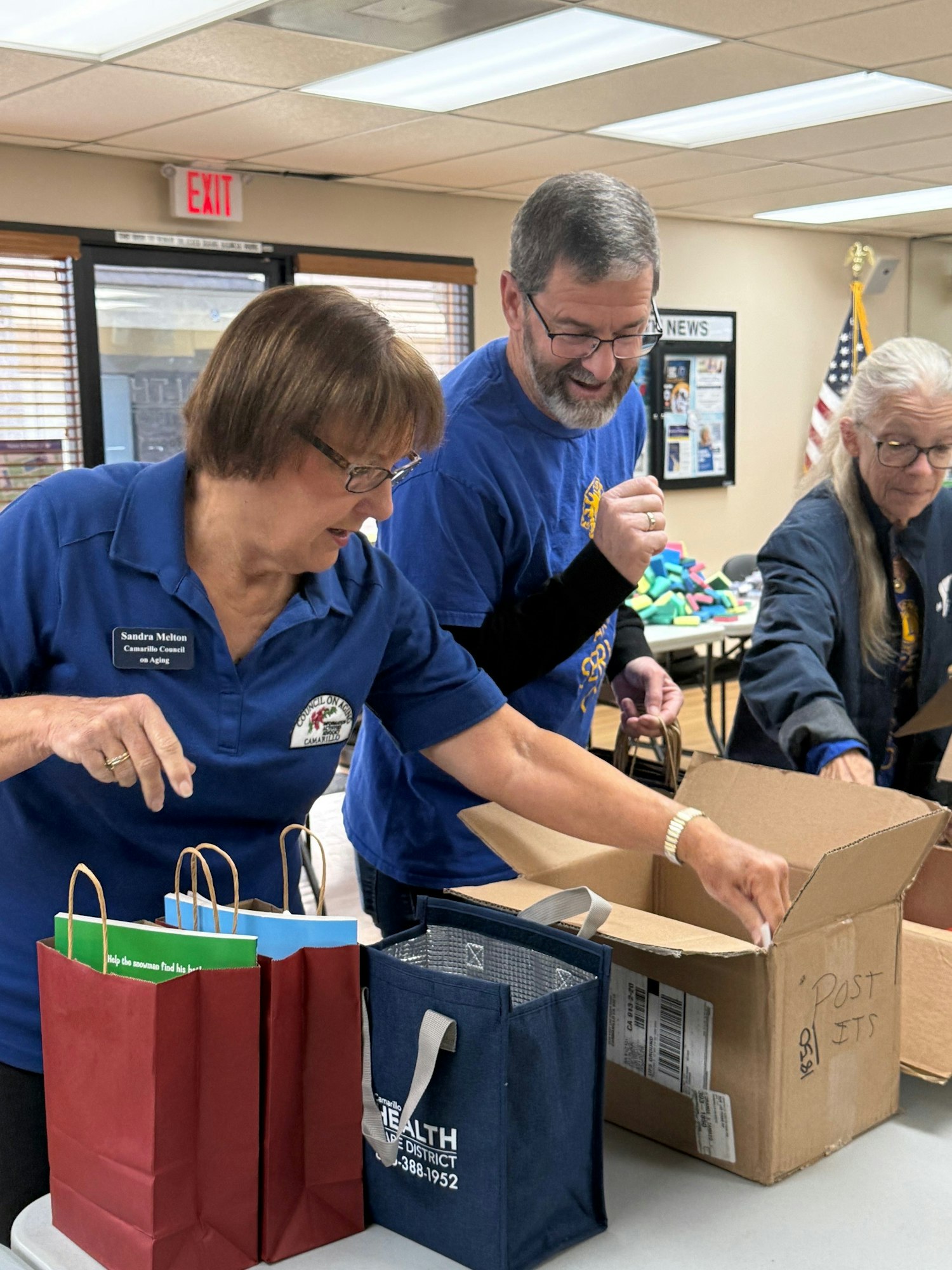 People packing items into gift bags on a table indoors, with boxes and an American flag in the background.
