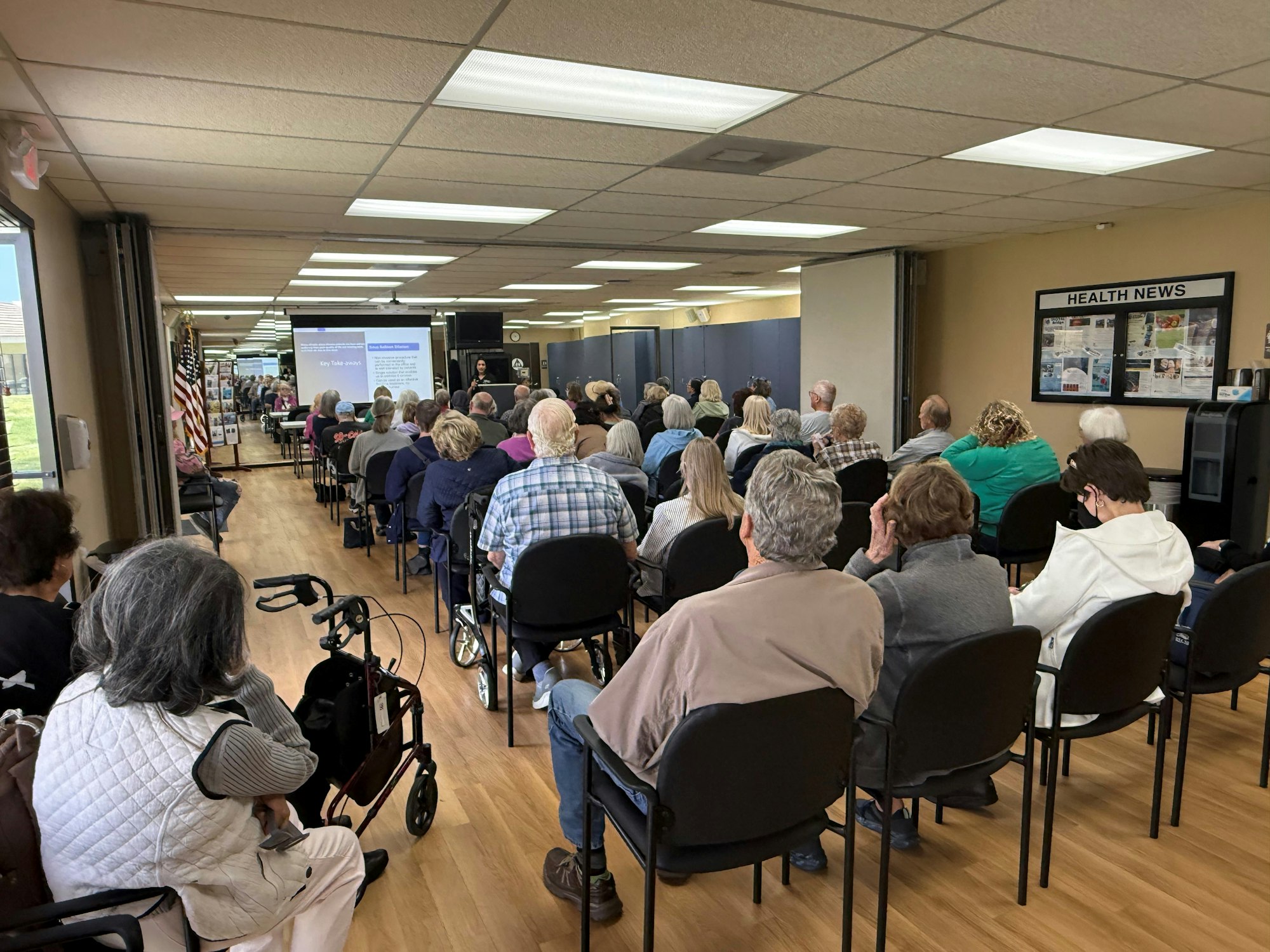 A group of people seated in a meeting room attending a presentation with a screen at the front displaying slides.