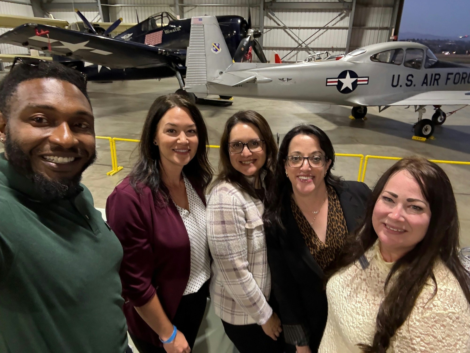 A group of five people smiling for a selfie in front of U.S. Air Force planes inside a hangar.