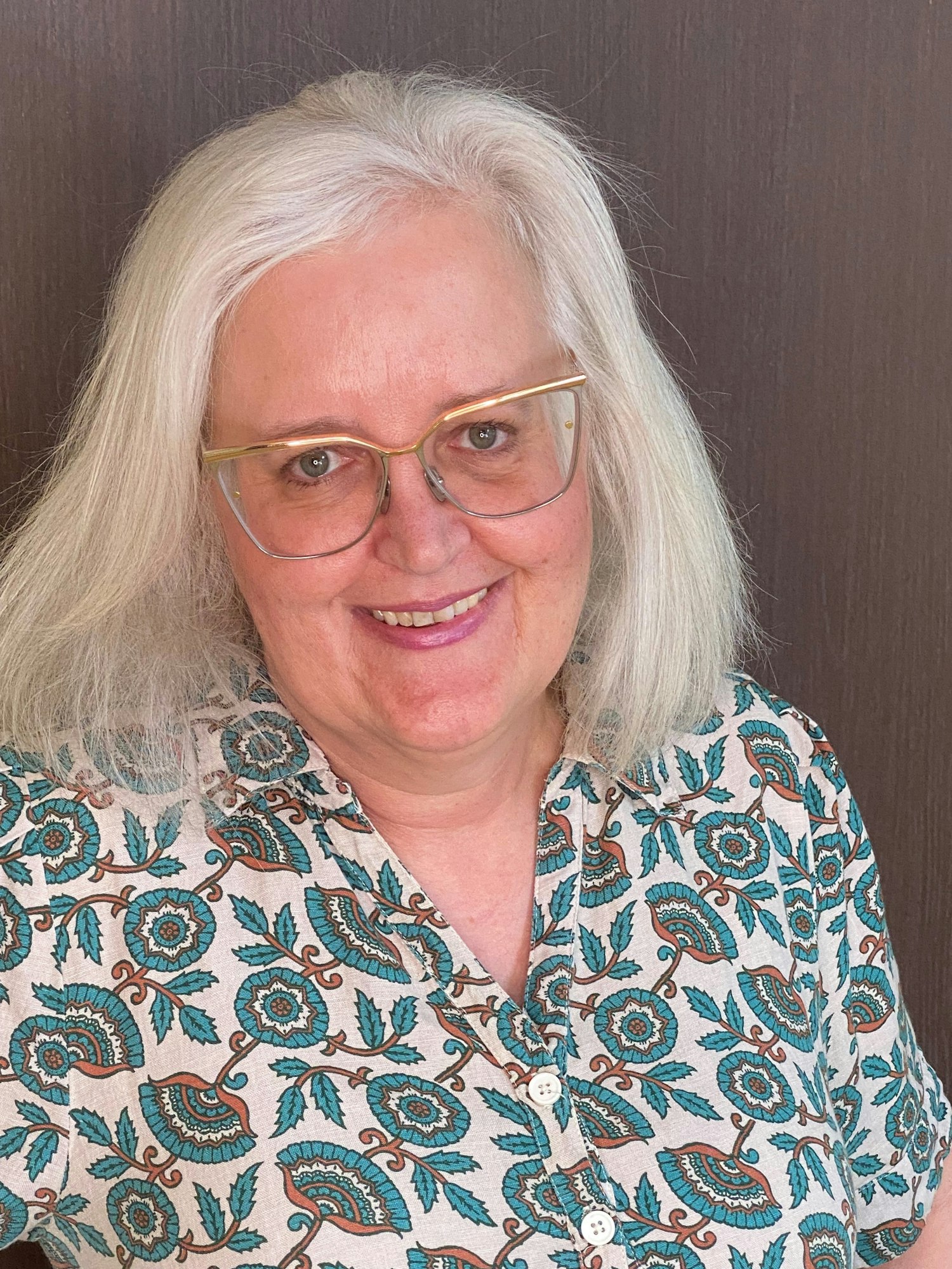 Smiling woman with white hair, glasses, and paisley patterned shirt against a dark background.