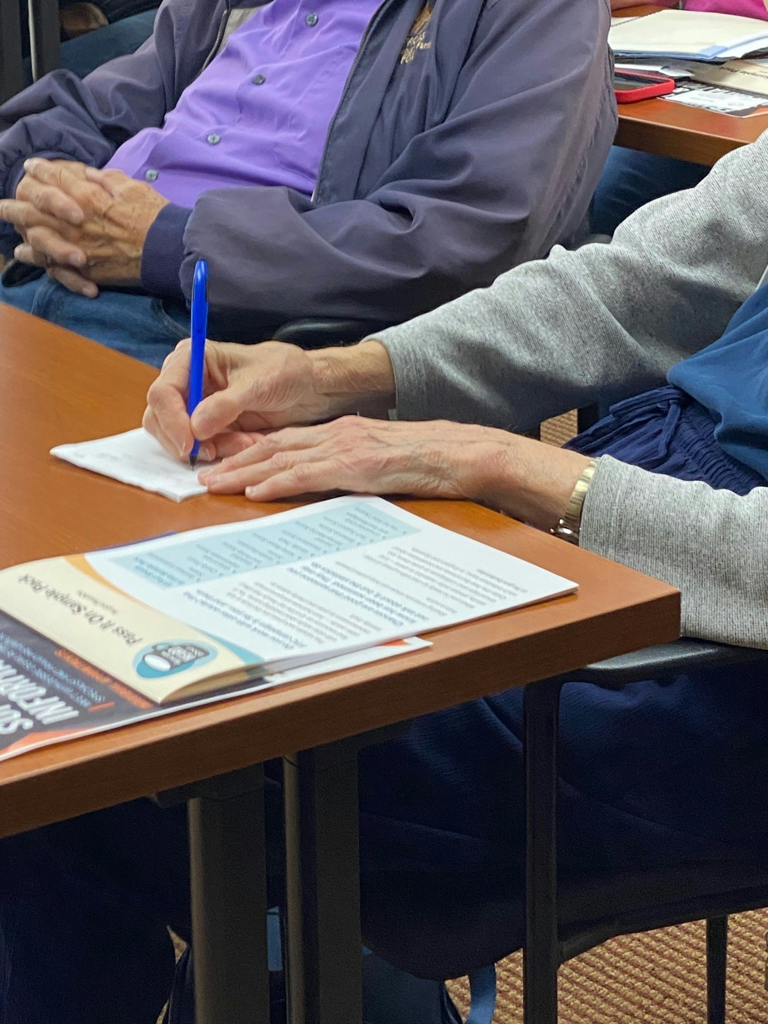 A person is writing on a notepad, with another person seated nearby, and a pamphlet on a table.