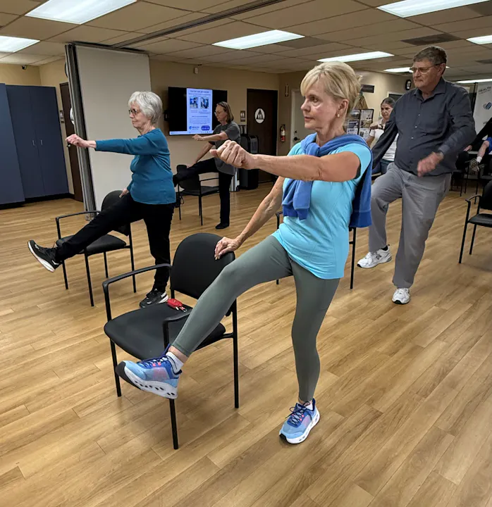 A group of people exercises in a room, using chairs for support while performing leg lifts and arm movements.