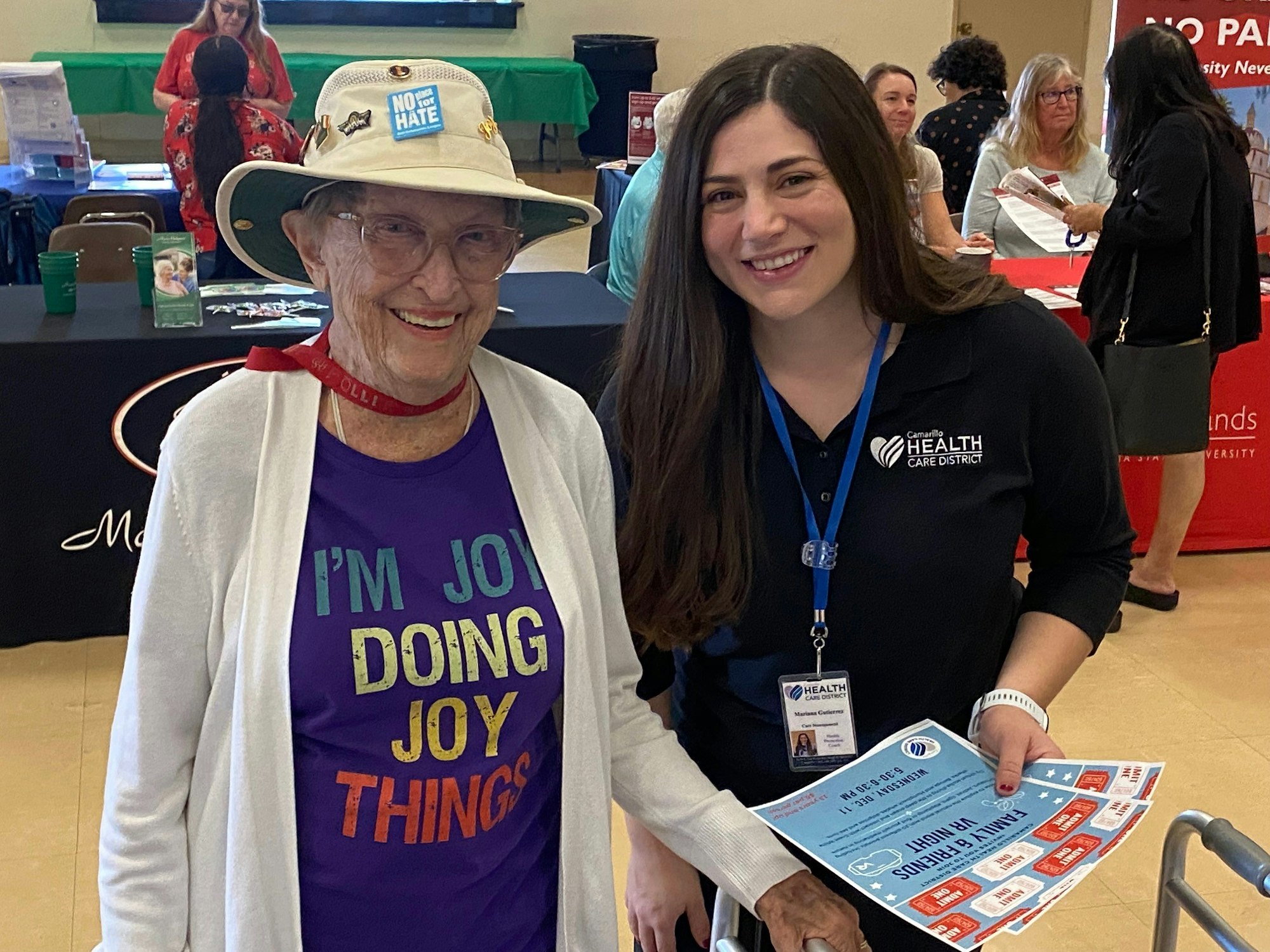 Two smiling women, one elderly with a "NO HATE" hat and "JOY" shirt, and a younger one with a badge, both holding pamphlets.