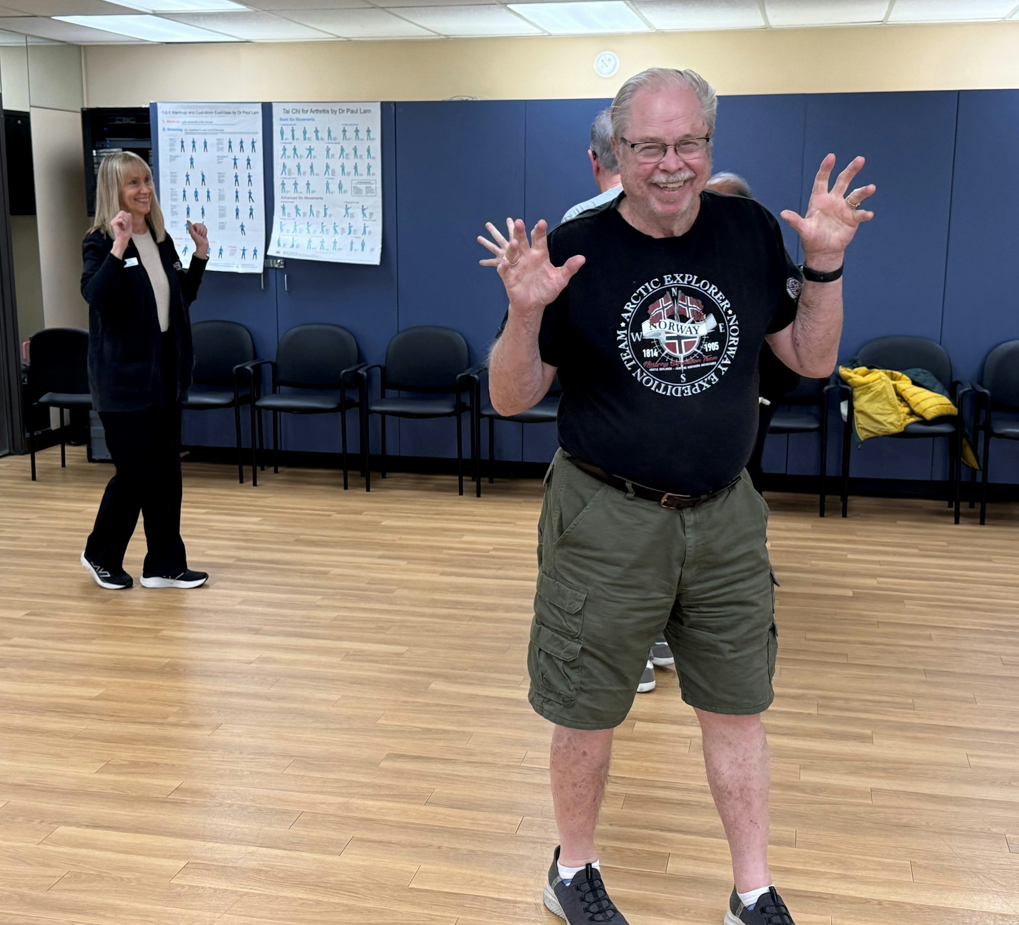 A man playfully poses with claw-like hands, while a woman in the background smiles, all in a spacious indoor setting.