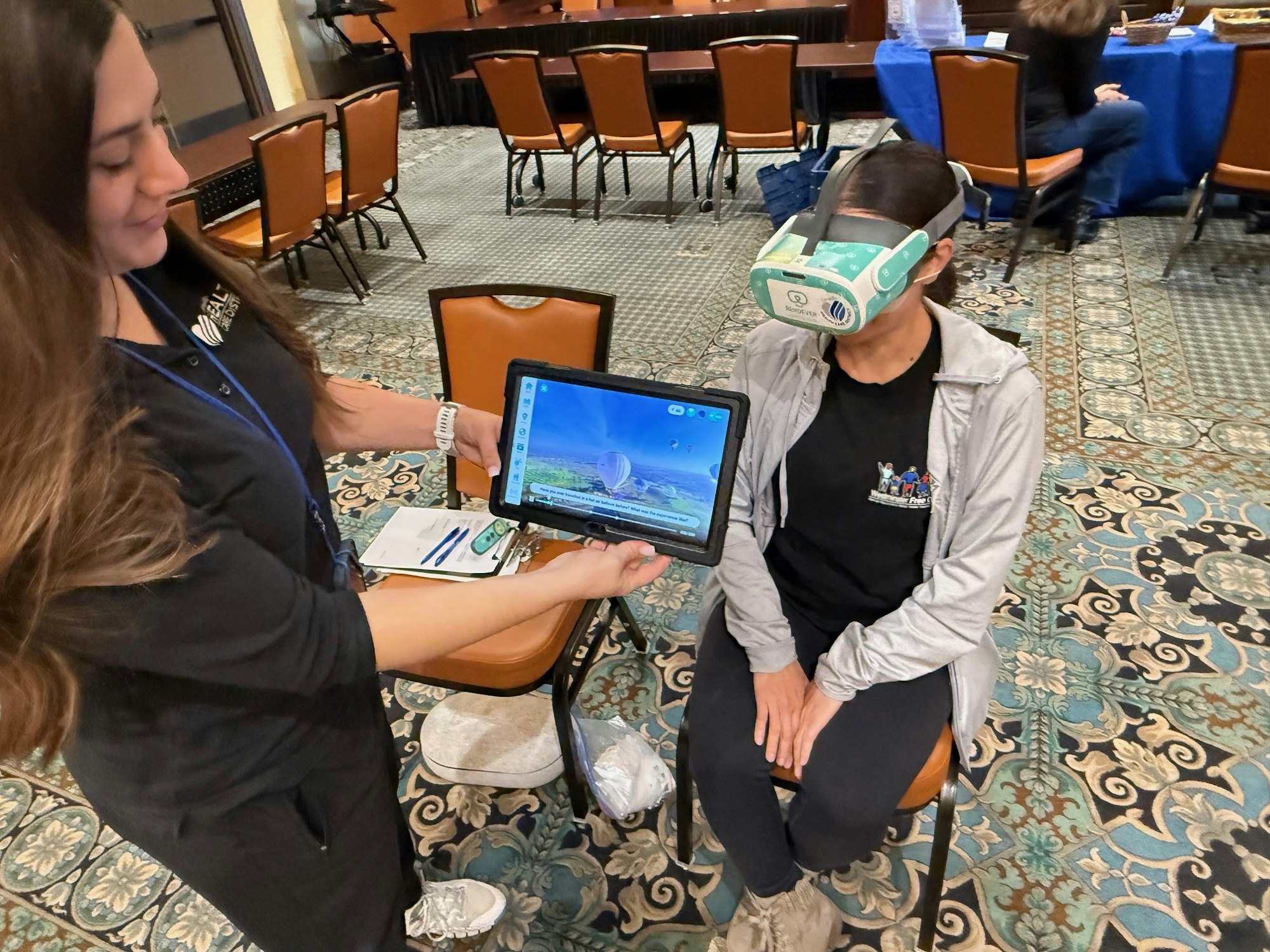 A person wearing a VR headset, sitting, while another holds a tablet displaying a landscape.