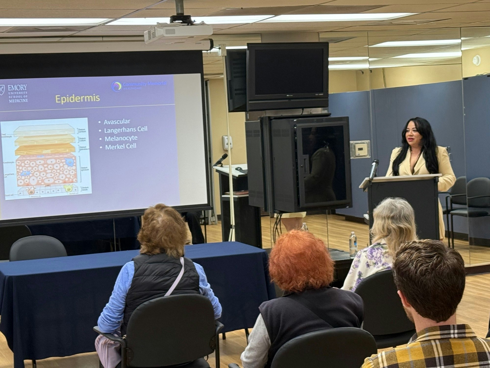 A woman presents a lecture on the epidermis, with a slide and audience in attendance.