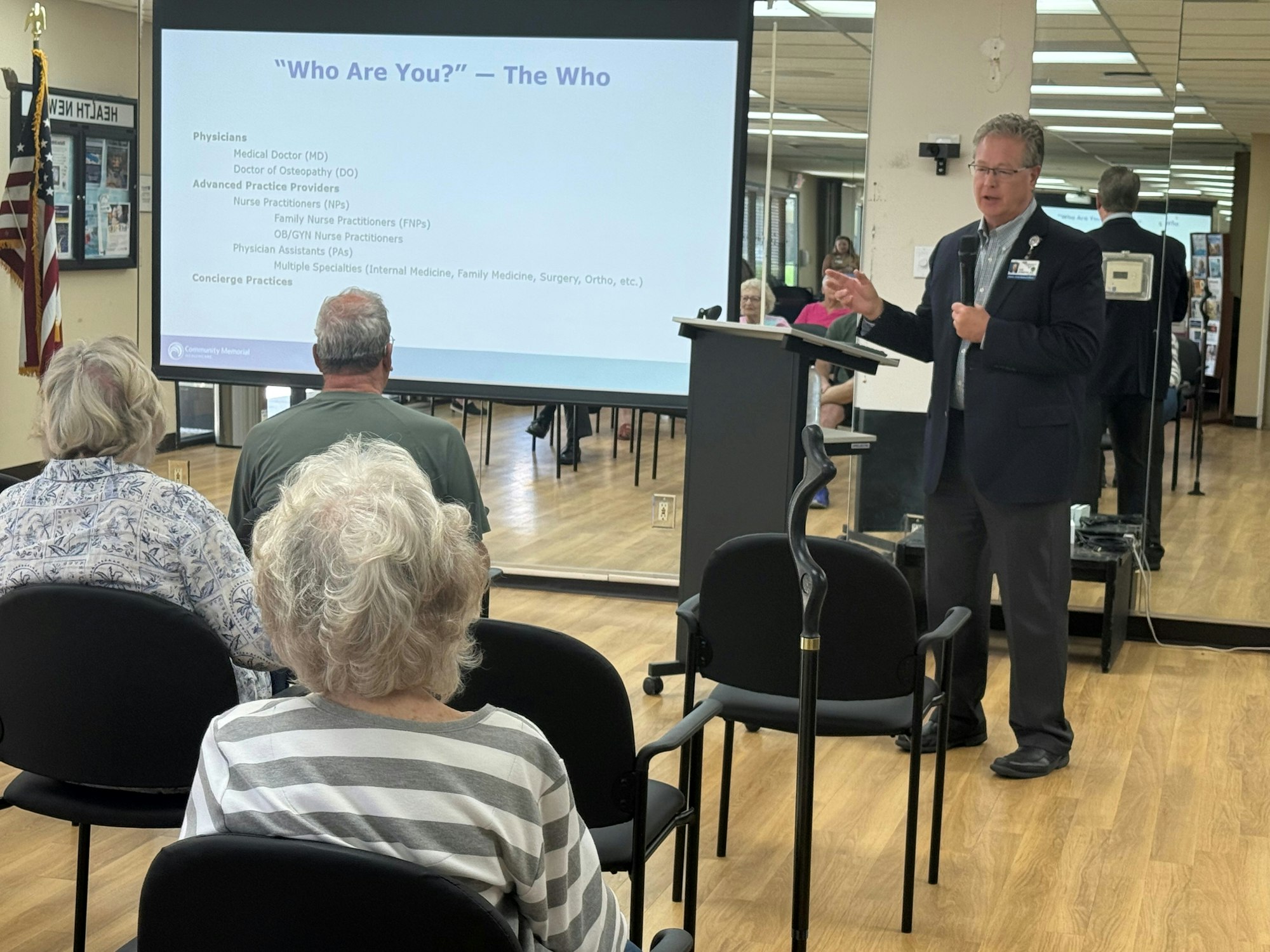 A speaker presents a slide titled "Who Are You?" to an audience in a meeting room.