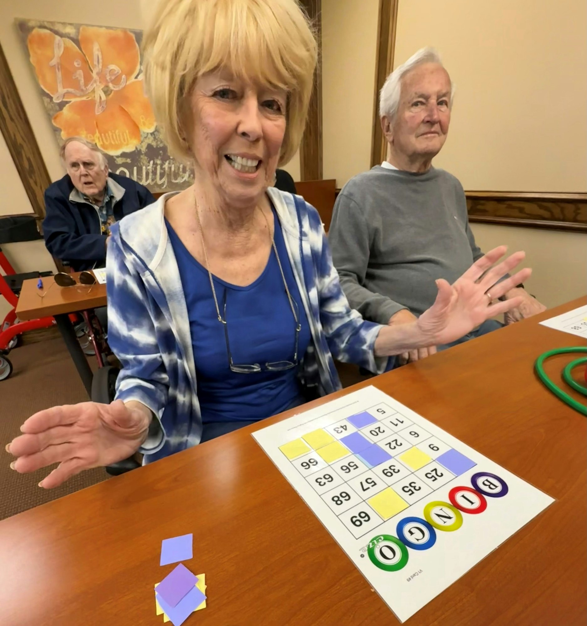 A smiling woman plays bingo at a table with two other people, showing her game card and colorful tiles.