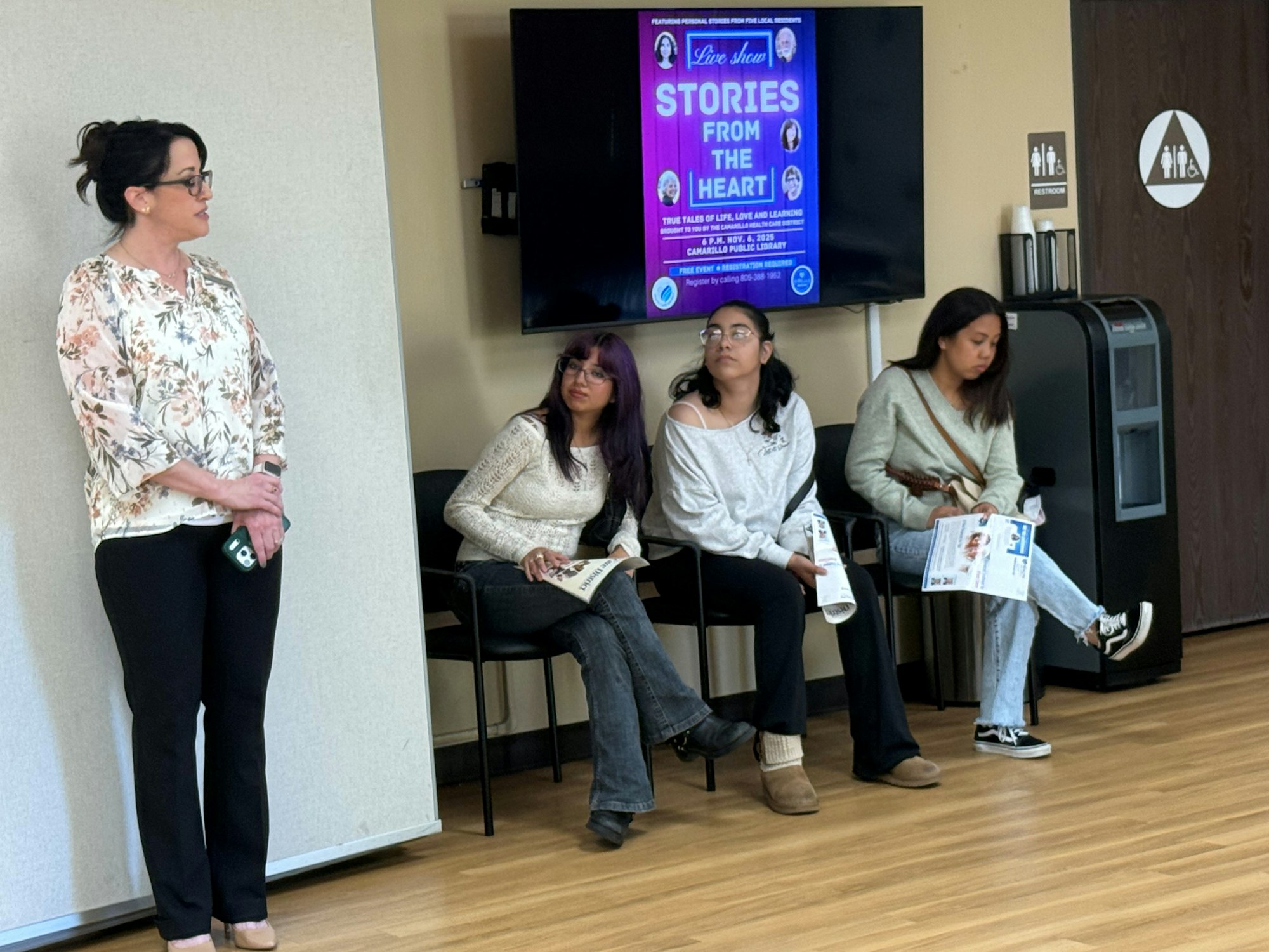 A woman stands speaking next to three seated people holding papers, with a "Stories from the Heart" event poster on a screen.