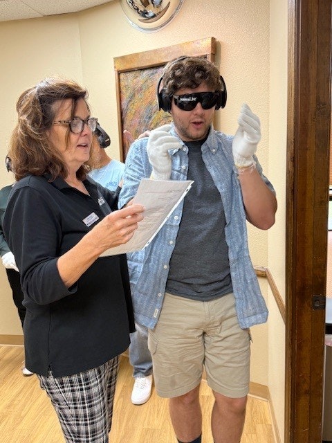 A man wearing sensory impairment goggles, gloves, and headphones listens to a woman holding a paper.
