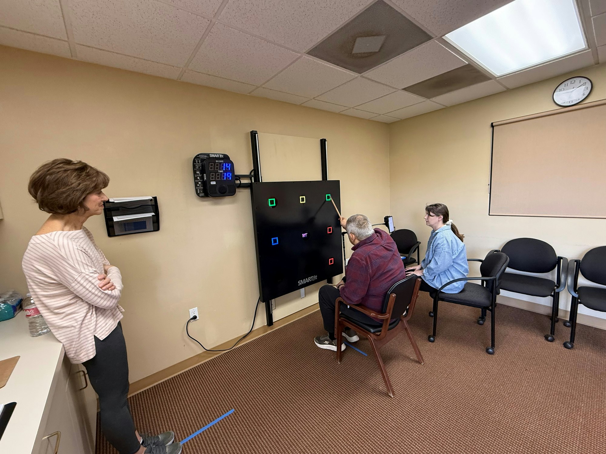 A man interacts with a touchscreen activity while a woman observes, in a room with chairs and a clock on the wall.