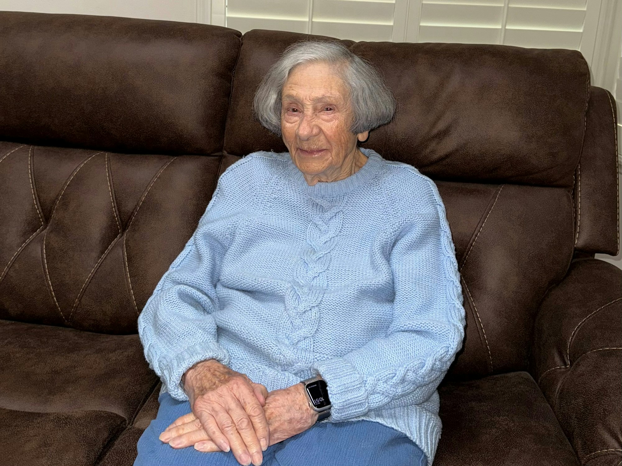 An elderly woman smiles while sitting on a brown couch, wearing a light blue sweater and resting her hands on her lap.