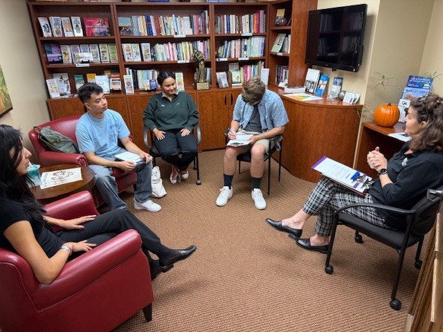 Five people sitting in a library-like office, engaged in discussion or meeting, surrounded by books and a pumpkin on the shelf.