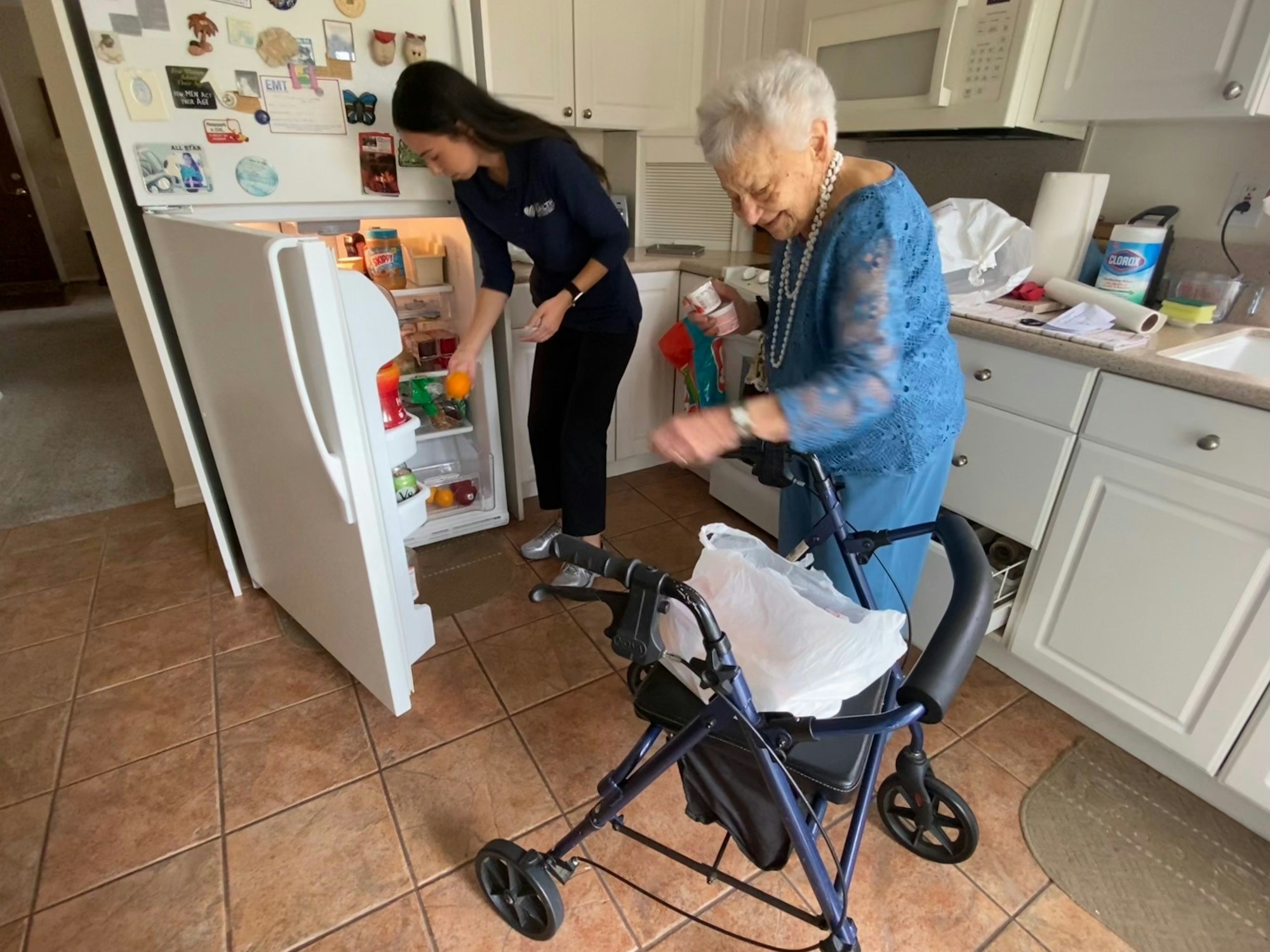 An elderly woman using a walker smiles near an open fridge, and a caregiver is organizing items inside it.