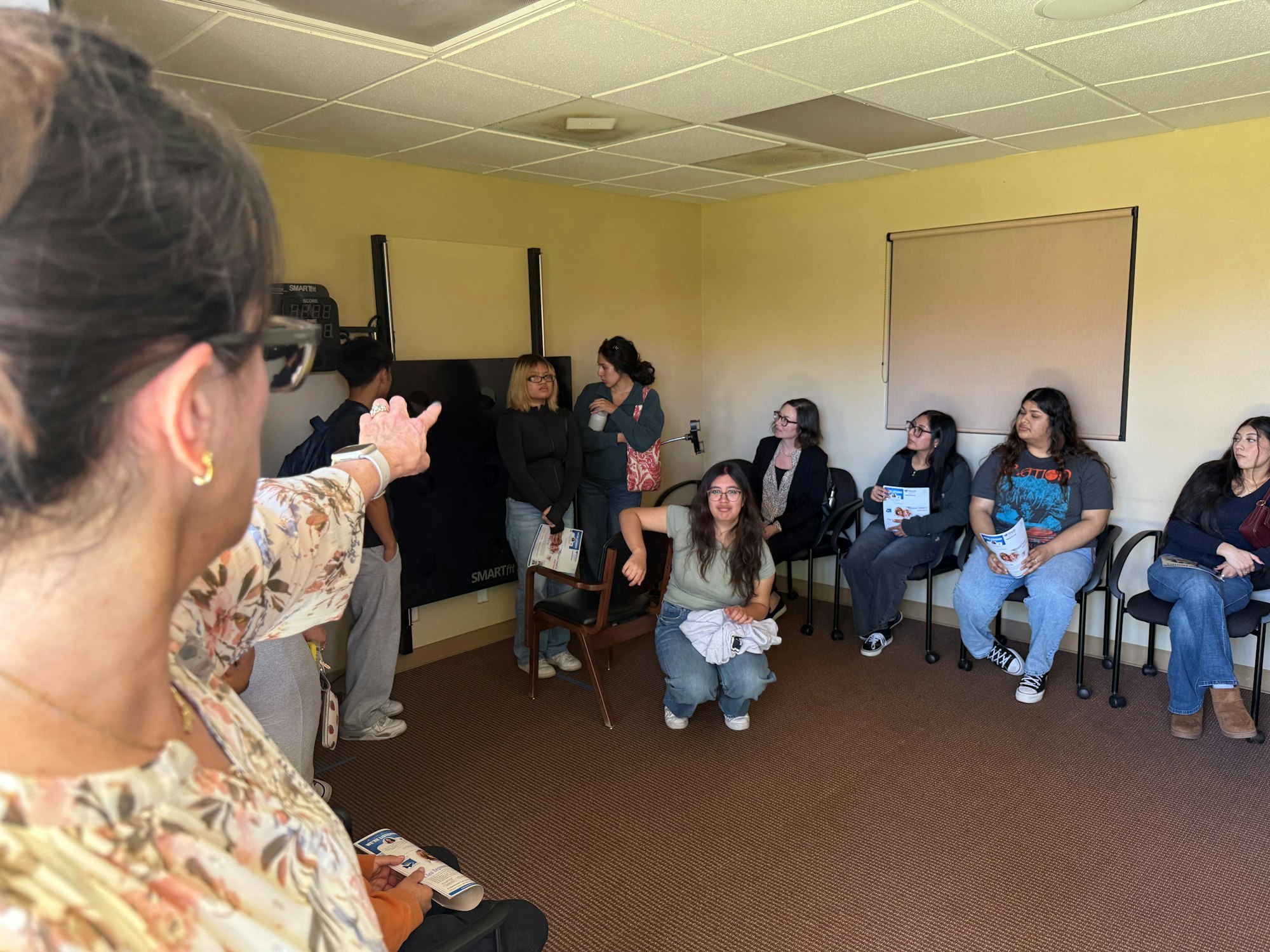 A woman gestures towards a group seated in a room. Some people hold booklets.