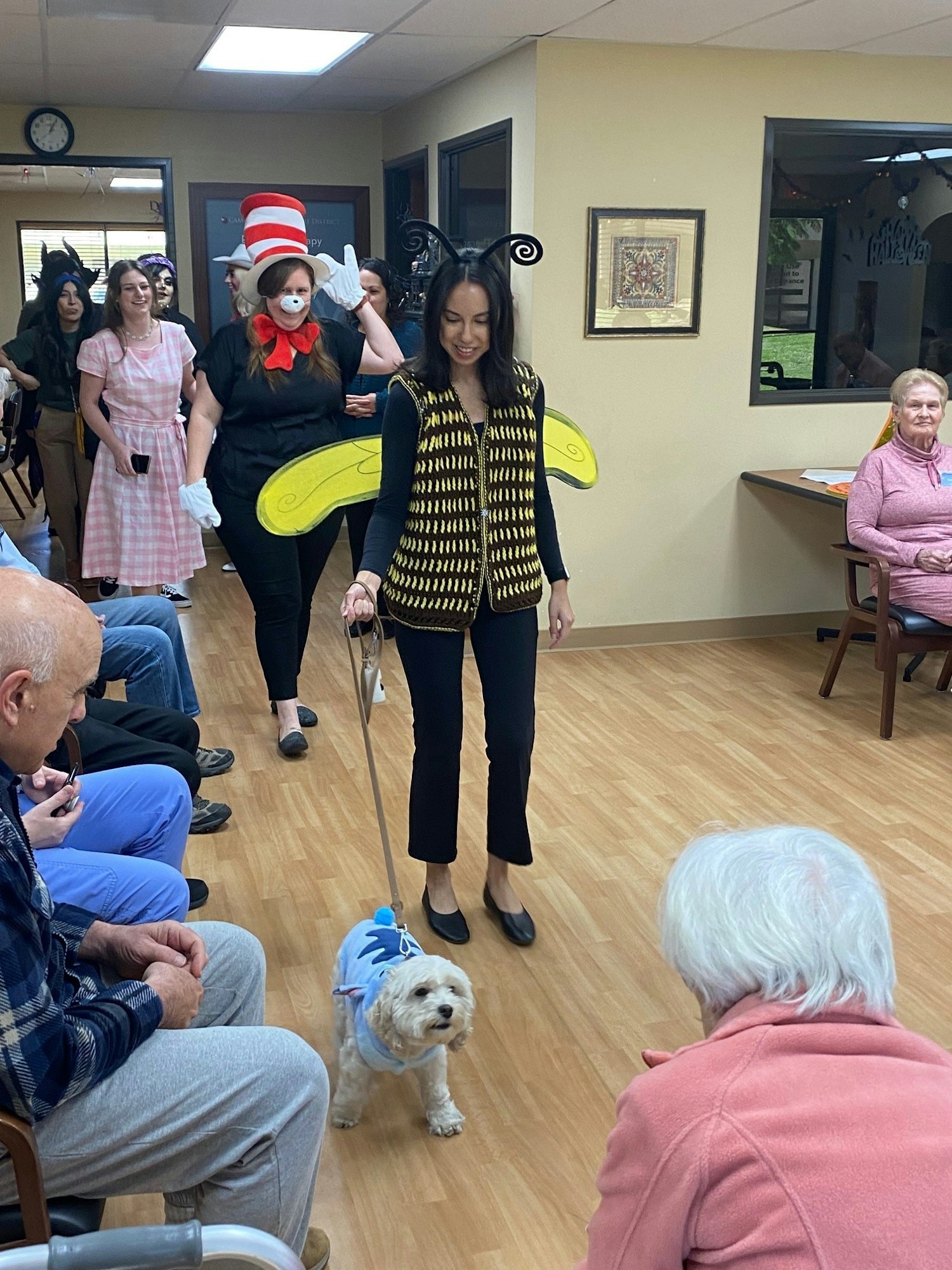 A woman dressed as a bee walks a small dog in a cape, surrounded by people in various costumes, with onlookers enjoying the scene.