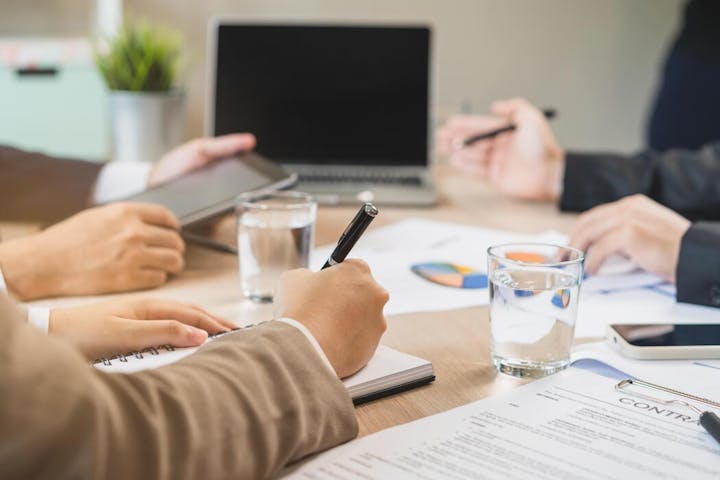 A busy meeting with people discussing materials, taking notes, and reviewing documents on a table, with a laptop and water glasses.