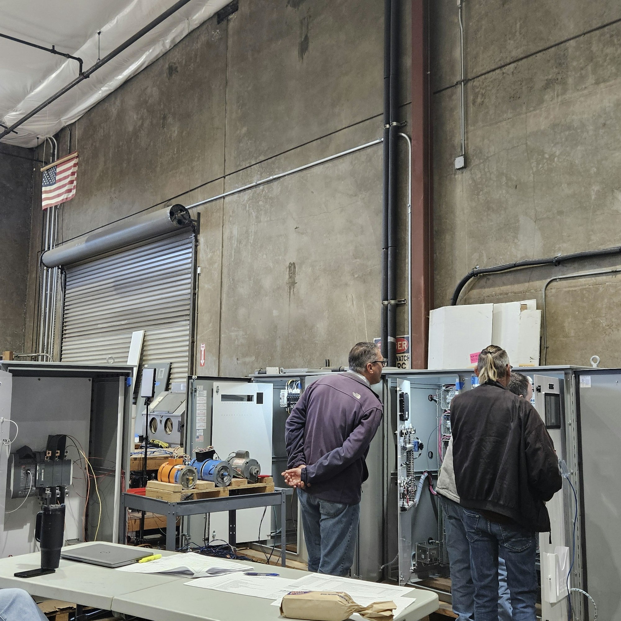Two individuals are observing equipment in a workshop, with machinery and an American flag visible in the background.