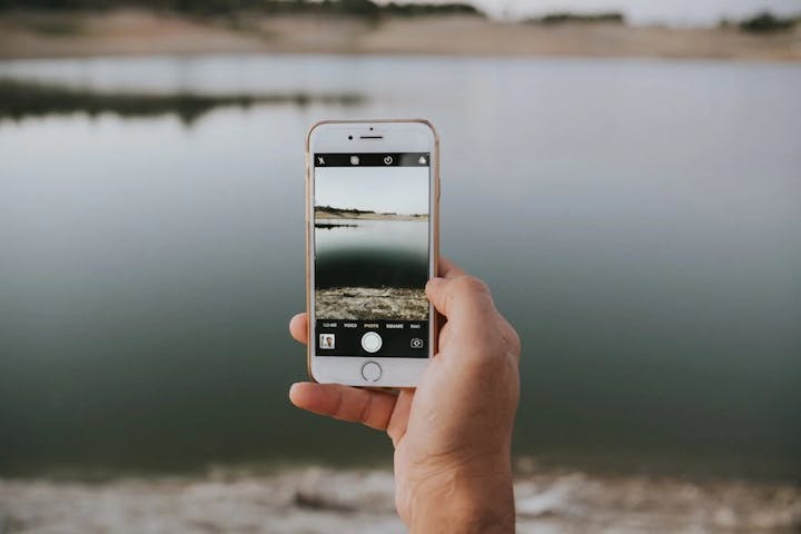 A person holding a smartphone, taking a photo of a calm body of water and scenic background.