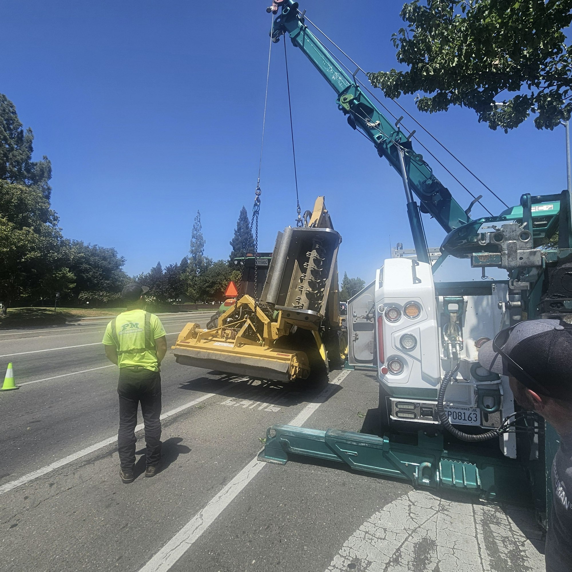 A crane lifting equipment onto a road, with a worker in a safety vest nearby and clear blue skies above.