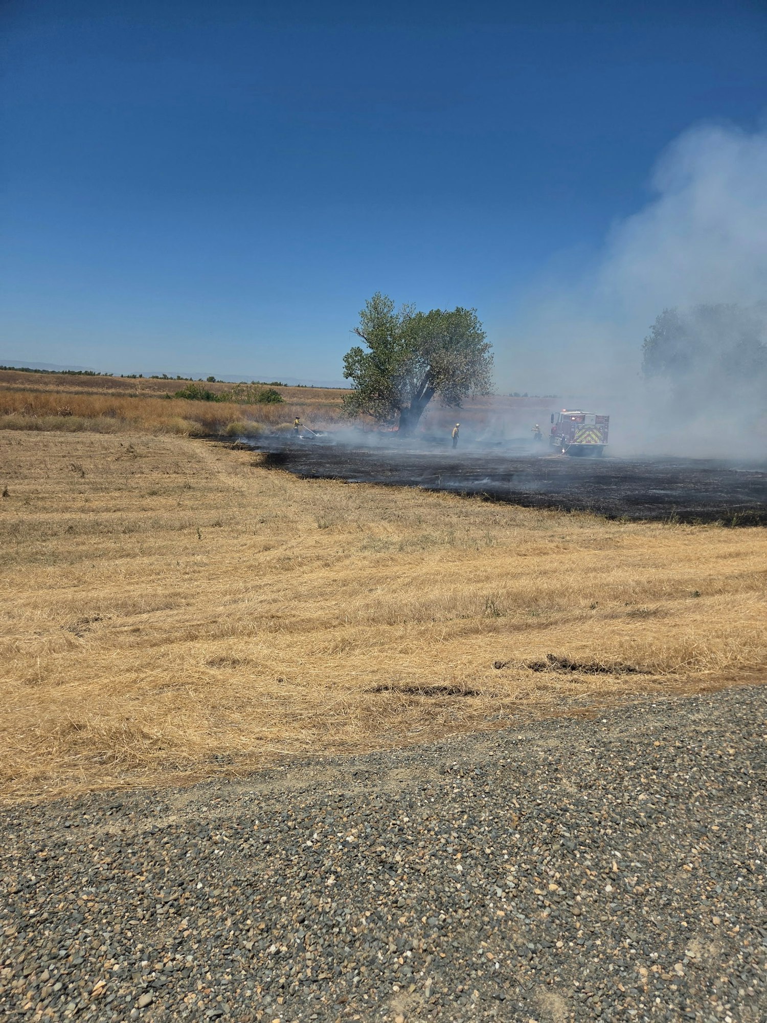 The image shows firefighters working to control a small grass fire in a field, with smoke rising against a blue sky.