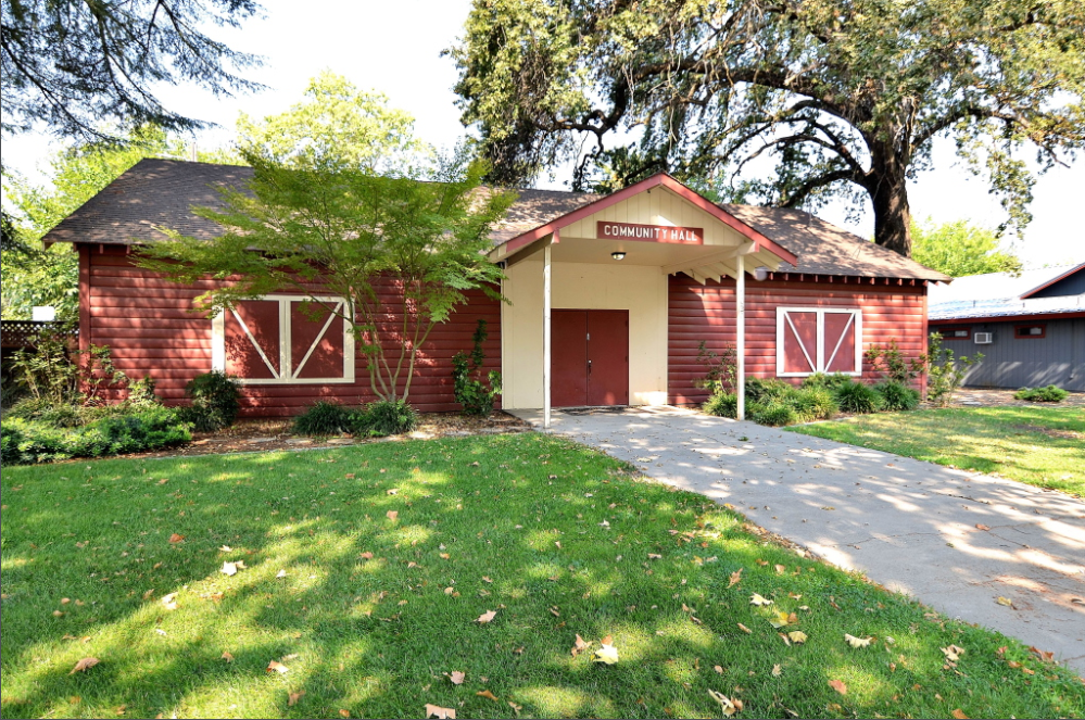 A red community hall building surrounded by greenery and trees, with a pathway leading to its entrance.