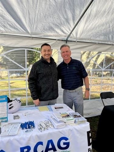 Two men stand smiling under a tent behind a table with brochures and pens, featuring an BCAG sign.